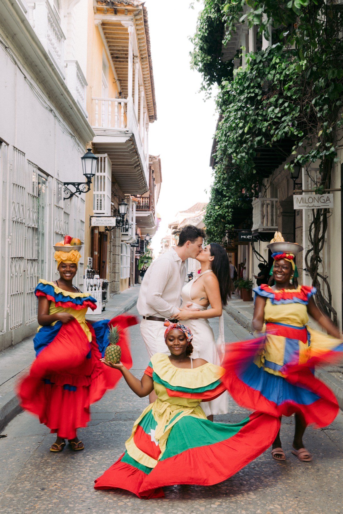 Couple kissing on a colorful Cartagena street, surrounded by palenqueras in traditional colorful dress